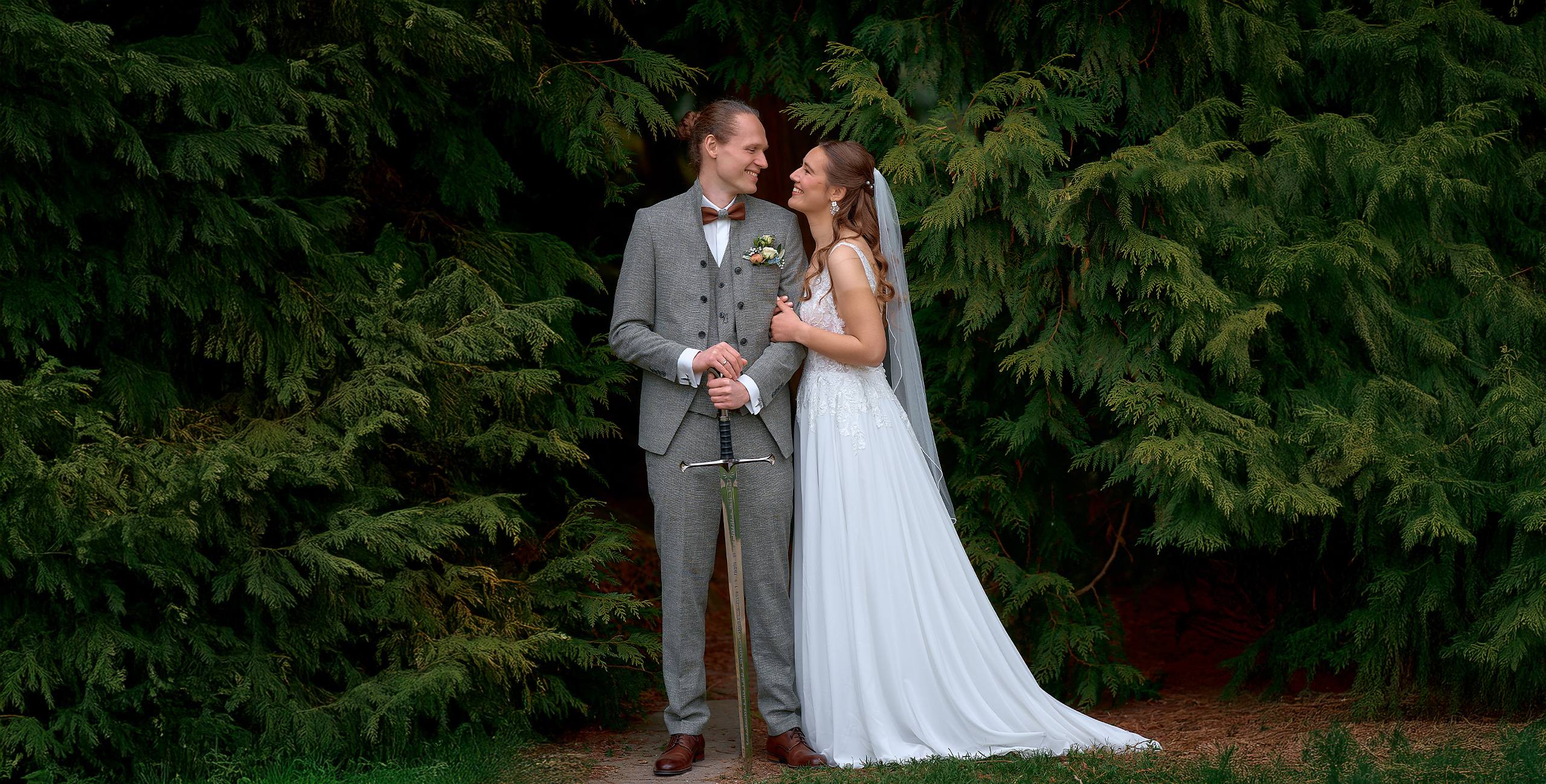 Ein Brautpaar steht eng beieinander vor dichtem, dunkelgrünem Nadelbaum-Gehölz und blickt sich lächelnd an. Die Braut trägt ein langes, weißes Kleid mit Schleier und legt liebevoll ihren Arm an den Bräutigam. Dieser ist in einem grauen Anzug mit Fliege gekleidet und hält ein dekoratives Schwert vor sich. Die Szene wirkt ruhig und romantisch, eingebettet in die natürliche Waldkulisse.