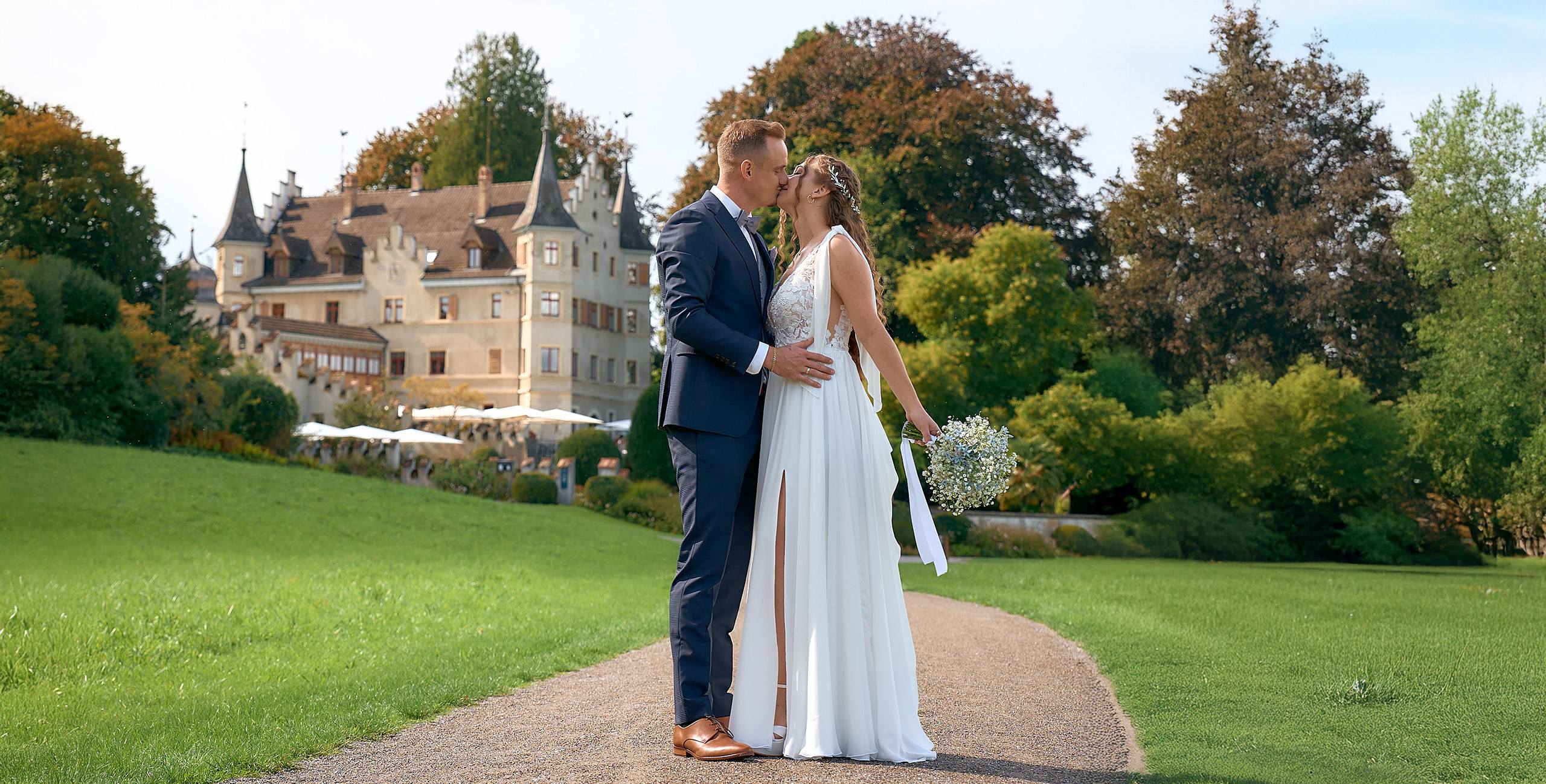 Ein Brautpaar steht auf einem geschwungenen Kiesweg in einem weitläufigen Park und küsst sich. Die Braut trägt ein weißes, ärmelloses Kleid mit Schlitz und hält einen kleinen Blumenstrauß, während der Bräutigam einen dunklen Anzug mit braunen Schuhen trägt. Im Hintergrund ist ein großes, schlossähnliches Gebäude mit Türmen zu sehen, umgeben von grünen Wiesen, Bäumen und herbstlich gefärbtem Laub.